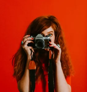 Portrait d'une photographe, une fille aux cheveux roux tenant un appareil photo sur fond rouge