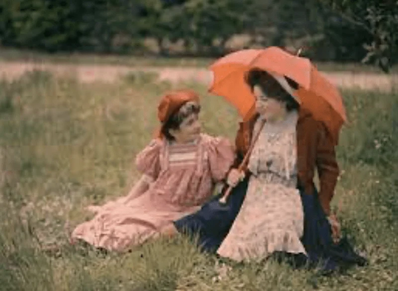 Photography of a woman holding a red umbrella and a girl by her side sitting in the middle of a green field, from 1910