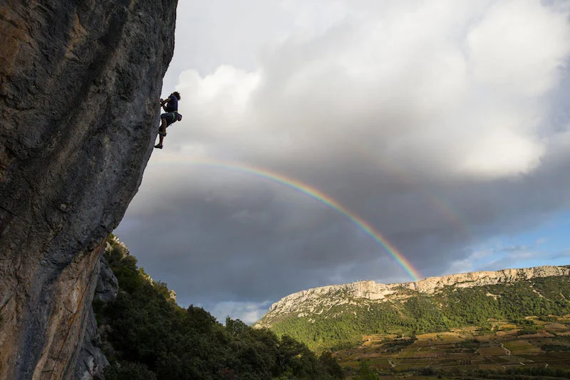 Photographie d'un homme escaladant une montagne avec un arc-en-ciel dans le ciel au Bousquet
