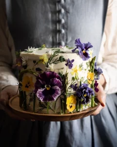 Photographie d'une personne tenant un gâteau couvert de fleurs jaunes et violettes, prise par Aimee Twigger, photographe culinaire