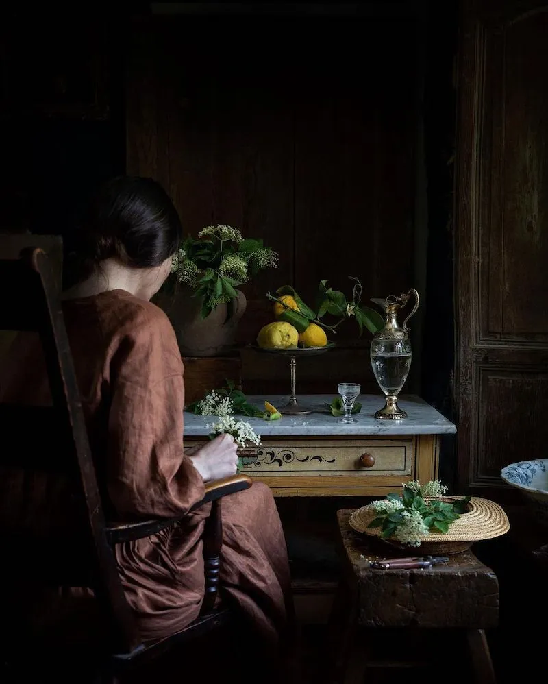 Photography of a woman looking to little flowers in front of a table with a bowl of lemons and a a pitcher of water